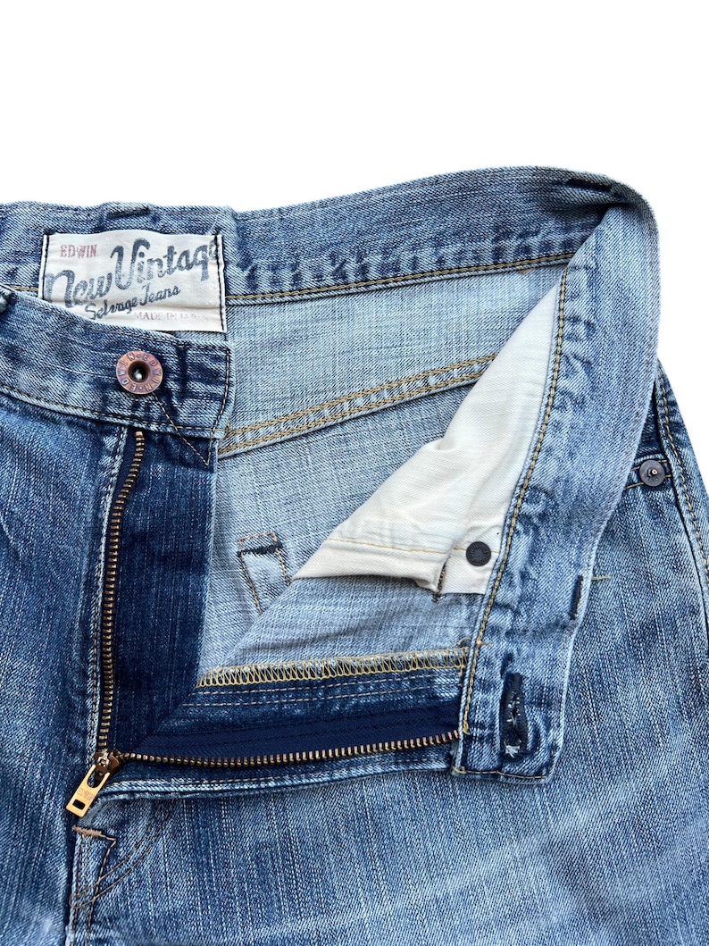 Pu&ograve; includere: Primo piano di jeans blu in denim con una cerniera in ottone parzialmente aperta. I jeans hanno una chiusura con bottone e una piccola etichetta con la scritta "New Vintage". Il denim ha un aspetto sbiadito.