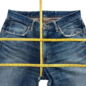 Puede incluir: Primer plano de unos vaqueros azules con una cinta m&eacute;trica amarilla colocada sobre la cintura y las piernas. Los vaqueros tienen un dise&ntilde;o cl&aacute;sico de cinco bolsillos y muestran signos de desgaste. La cinta m&eacute;trica est&aacute; en cent&iacute;metros.