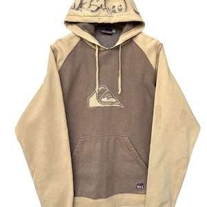 May include: A tan and brown hoodie with a kangaroo pocket and drawstring. The hood features embroidered text, and the chest displays a logo. The sleeves and hood are tan, while the body is brown. A casual, comfortable garment.