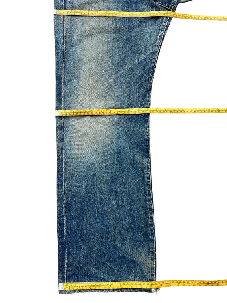 May include: Close-up of blue denim jeans with yellow measuring tape. The jeans are laid flat, showing the waist, thigh, and length measurements. The denim has a faded, worn appearance.