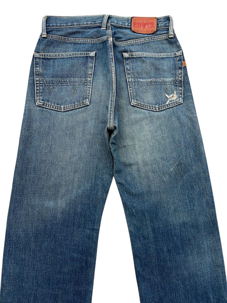 Puede incluir: Vaqueros de mezclilla azul descolorido con un dise&ntilde;o cl&aacute;sico de cinco bolsillos. Los vaqueros presentan un aspecto desgastado con un parche desgastado en el bolsillo trasero y un parche de marca de cuero rojo. La etiqueta de la marca EDWIN es visible.