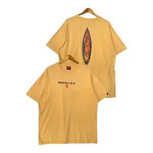 May include: Mustard yellow Oakley t-shirts. One shirt features the Oakley logo in brown and red on the front. The other shirt has a surfboard graphic with flames on the back. Both are short-sleeved.
