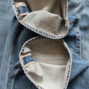 May include: Close-up of blue denim jeans with the cuffs turned up. The inside of the cuffs is a lighter shade of denim, with orange stitching visible along the edges. The jeans have a classic five-pocket design.