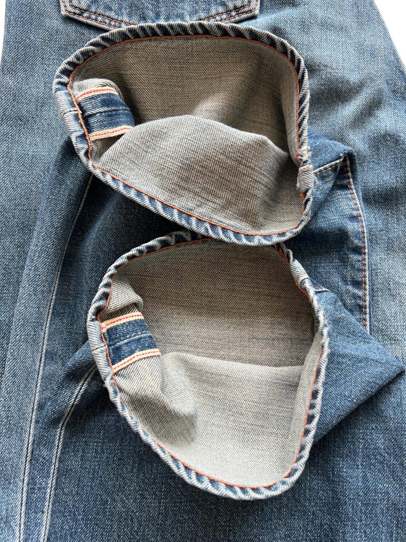 May include: Close-up of blue denim jeans with the cuffs turned up. The inside of the cuffs is a lighter shade of denim, with orange stitching visible along the edges. The jeans have a classic five-pocket design.