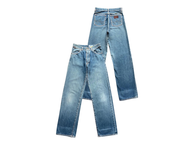 May include: Two pairs of light blue denim jeans. One pair is laid flat, showing the front, while the other is angled, displaying the back pockets and a brown leather patch. The jeans have a classic, straight-leg fit.