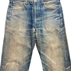 May include: Faded blue denim jeans with a button fly and classic five-pocket design. The jeans show signs of wear and tear, with faded patches and distressed areas. The jeans have a straight leg cut and a medium rise.