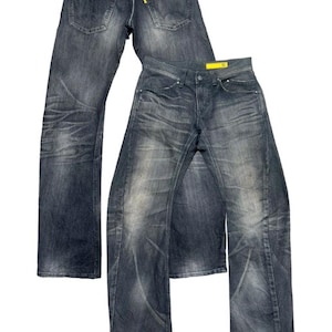 Può includere: Due paia di jeans in denim lavaggio scuro. Un paio è sovrapposto all'altro. I jeans hanno un aspetto sbiadito con segni di usura visibili. Le tasche posteriori e un'etichetta gialla sono visibili.