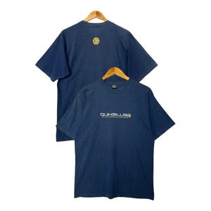 May include: Two navy blue t-shirts hanging on wooden hangers. The front shirt has the word "QUIKSILVER" in white and yellow. The back shirt has a yellow circular logo. Both shirts are short-sleeved.