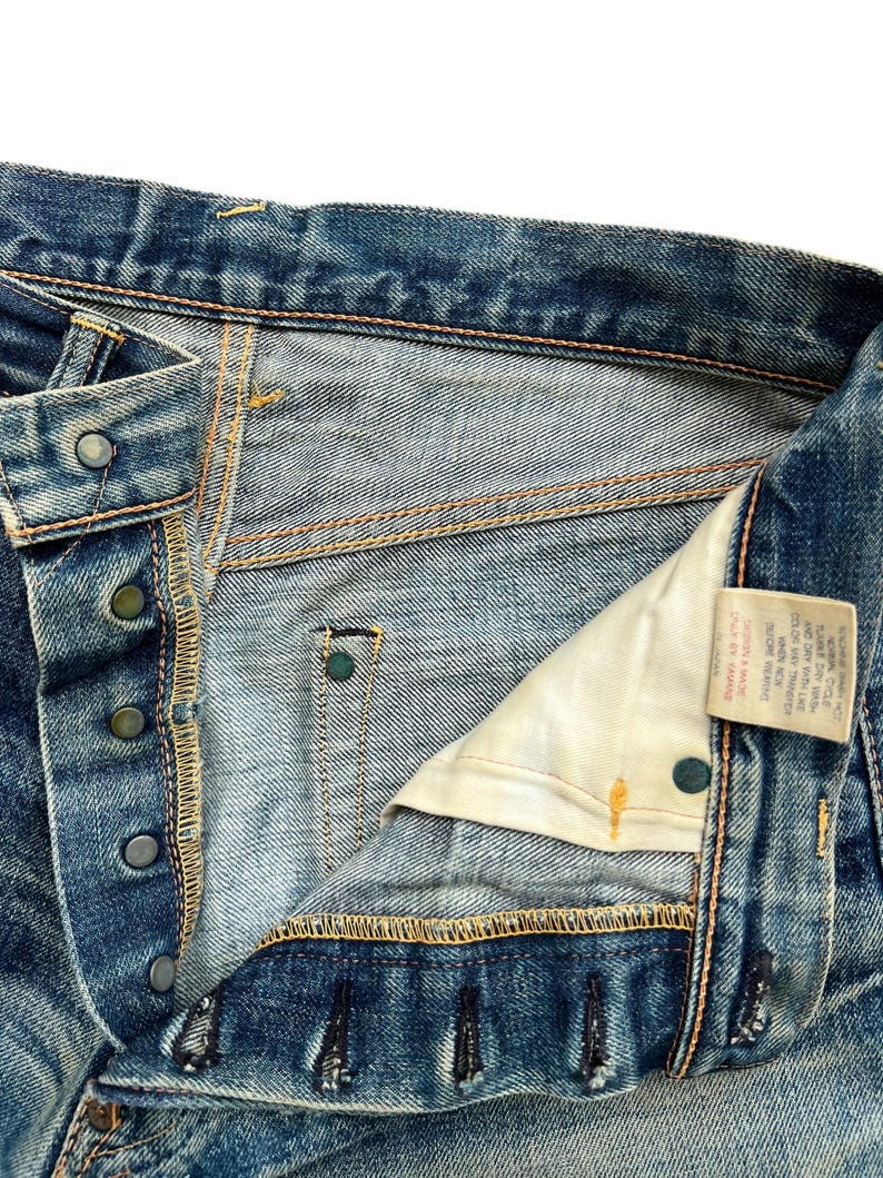 May include: Close-up of blue denim jeans with a button fly. The jeans show signs of wear and fading, with visible stitching and metal button details. A small tag is visible on the inside.