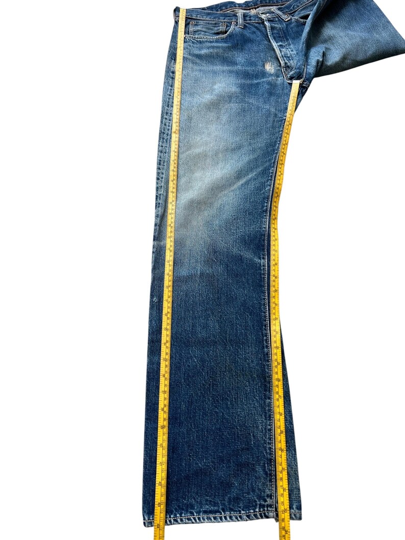 以下が含まれることがあります： ボタンフライの青いデニムジーンズ。黄色の巻尺で測定されています。ジーンズは使用感と色あせが見られます。巻尺はジーンズの長さを示しています。