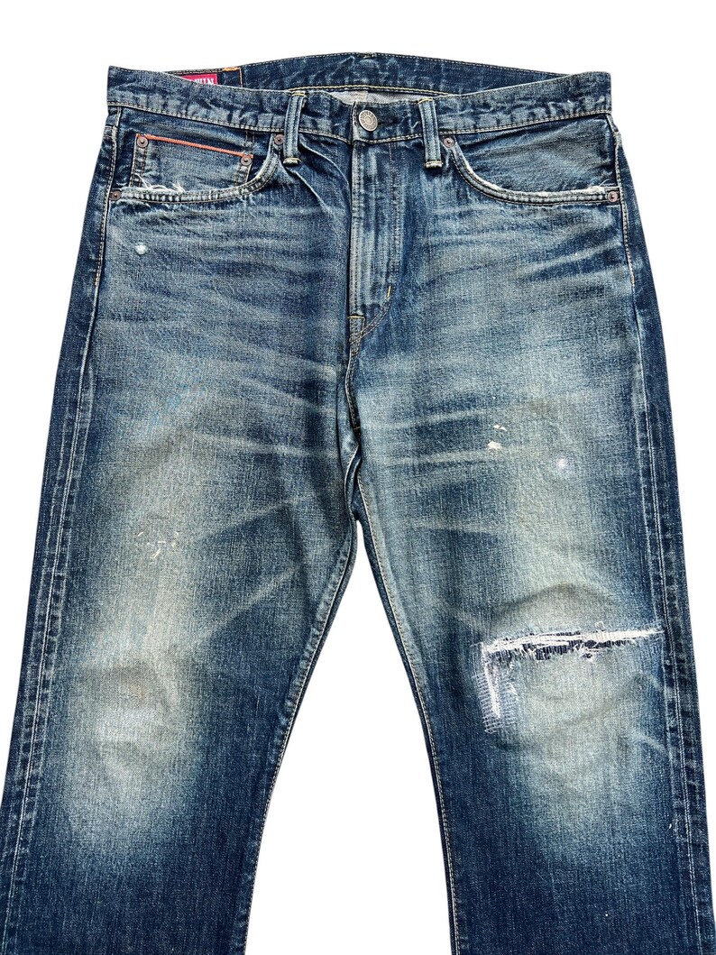 Puede incluir: Vaqueros de mezclilla azul descolorido con aspecto desgastado. Los vaqueros tienen cierre de bot&oacute;n, dise&ntilde;o cl&aacute;sico de cinco bolsillos y rodilla parcheada. La mezclilla tiene un aspecto desgastado con decoloraci&oacute;n clara y sutil desgaste.