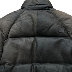 May include: Close-up of a black leather puffer jacket. The jacket features a high collar and a quilted design. The leather appears worn, with visible texture and variations in color. The stitching is visible, highlighting the panels of the jacket.