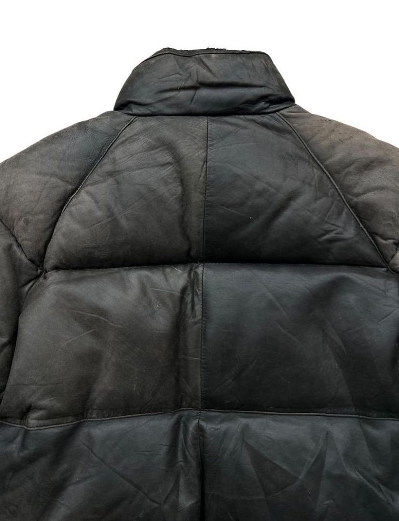 May include: Close-up of a black leather puffer jacket. The jacket features a high collar and a quilted design. The leather appears worn, with visible texture and variations in color. The stitching is visible, highlighting the panels of the jacket.