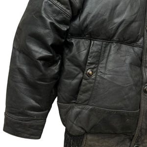 May include: Close-up of a black leather jacket with a quilted design. The jacket features a pocket with a button closure and a ribbed elastic hem. The leather has a worn, textured appearance.