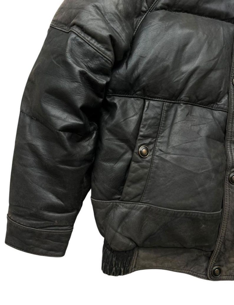 May include: Close-up of a black leather jacket with a quilted design. The jacket features a pocket with a button closure and a ribbed elastic hem. The leather has a worn, textured appearance.