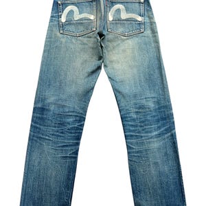 May include: Faded blue denim jeans with white curved designs on the back pockets. The jeans have a classic five-pocket design, a red leather patch, and a straight leg cut. The denim shows signs of wear and fading.