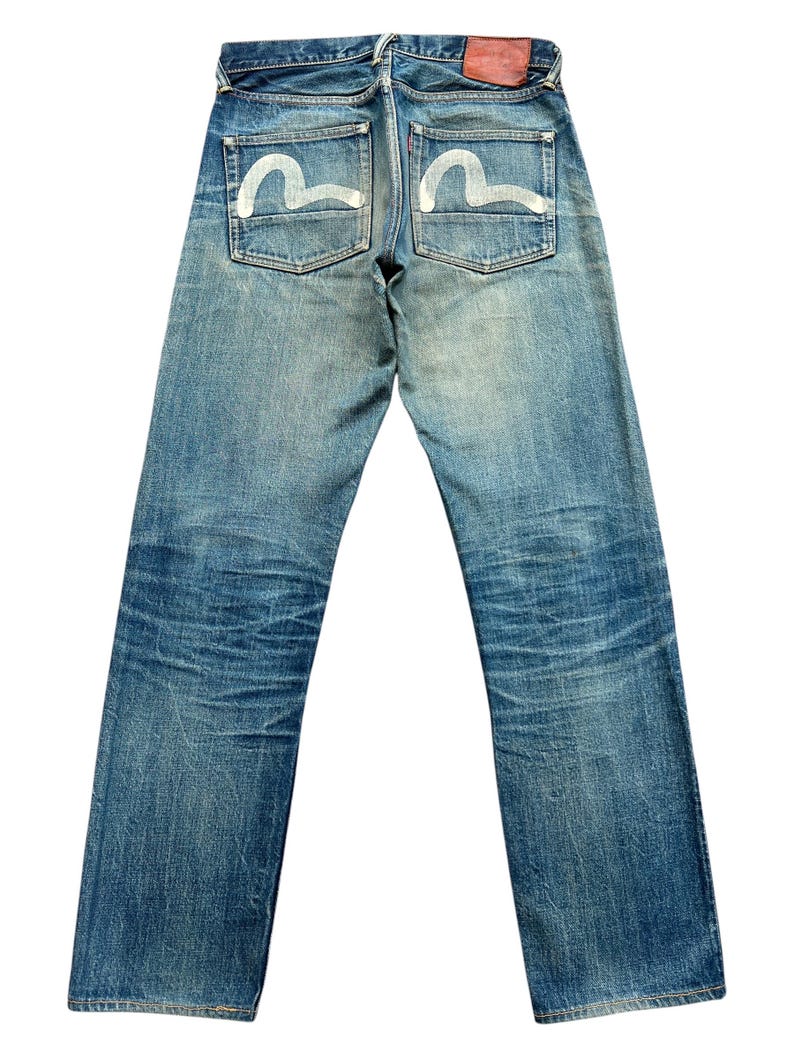 May include: Faded blue denim jeans with white curved designs on the back pockets. The jeans have a classic five-pocket design, a red leather patch, and a straight leg cut. The denim shows signs of wear and fading.