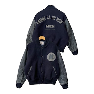 May include: Navy blue varsity jacket with gray leather sleeves. The jacket features the text "COMME ÇA DU MODE" on the back and a logo on the front. The jacket is button-down with a ribbed collar and cuffs.