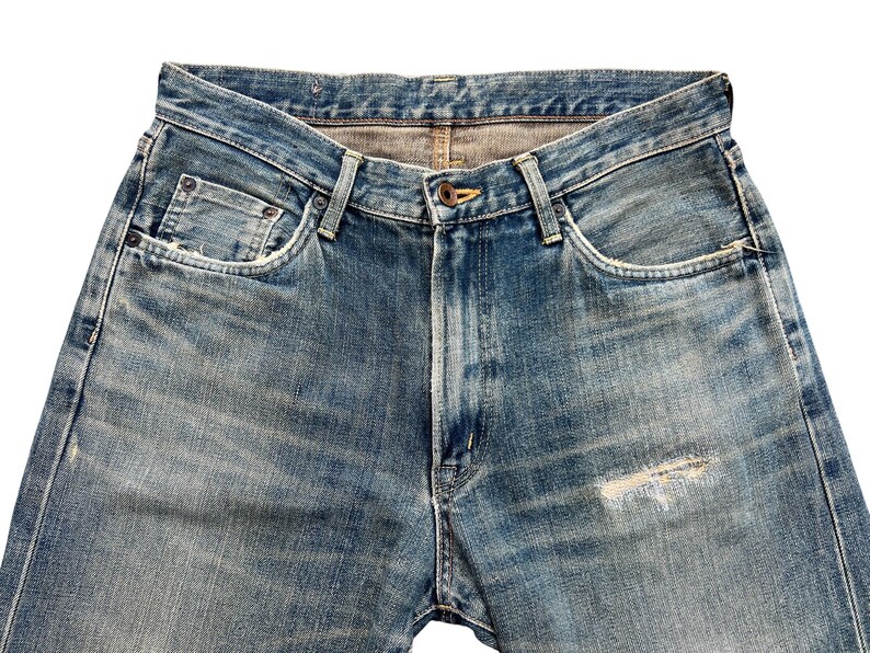 May include: Close-up of light blue denim jeans. The jeans have a classic five-pocket design, a button closure, and a distressed area on the front. The denim has a faded, worn look.
