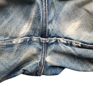 May include: Close-up of light blue denim jeans with distressed details. The image shows the crotch area of the jeans, with visible wear and tear, including faded patches and frayed edges. The stitching is visible.