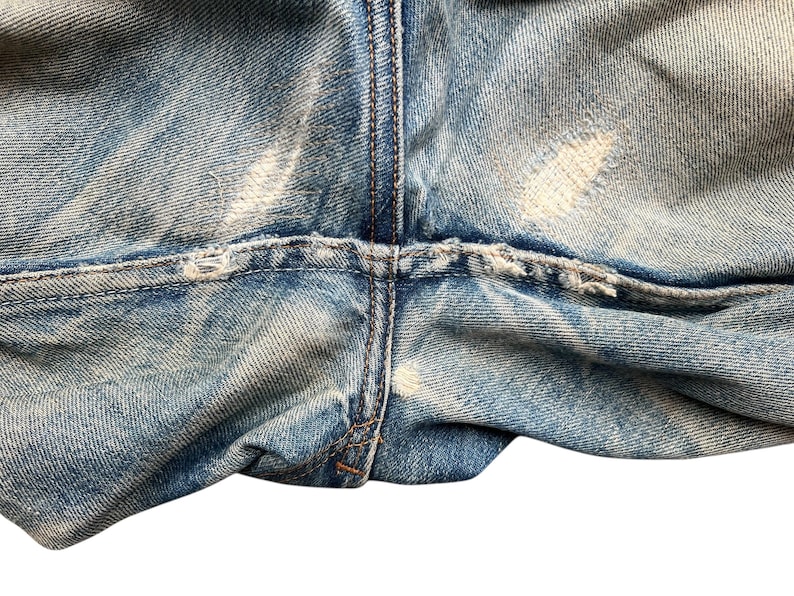 May include: Close-up of light blue denim jeans with distressed details. The image shows the crotch area of the jeans, with visible wear and tear, including faded patches and frayed edges. The stitching is visible.