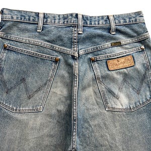 K&ouml;nnte beinhalten: Nahaufnahme der R&uuml;ckseite einer hellblauen Wrangler-Jeans. Die Jeans hat zwei Ges&auml;&szlig;taschen mit dem Wrangler-Logo auf der rechten Tasche. Die Jeans hat einen braunen Lederaufn&auml;her mit dem Wrangler-Logo.