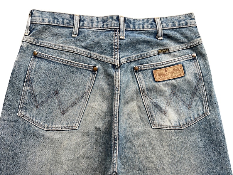 K&ouml;nnte beinhalten: Nahaufnahme der R&uuml;ckseite einer hellblauen Wrangler-Jeans. Die Jeans hat zwei Ges&auml;&szlig;taschen mit dem Wrangler-Logo auf der rechten Tasche. Die Jeans hat einen braunen Lederaufn&auml;her mit dem Wrangler-Logo.