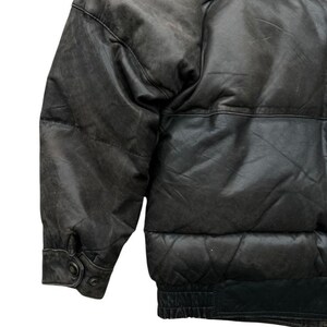 May include: A close-up of a black leather puffer jacket. The jacket has a quilted design and a ribbed hem. The sleeves have buttoned cuffs. The leather appears worn, with a matte finish. The jacket is a classic outerwear piece.