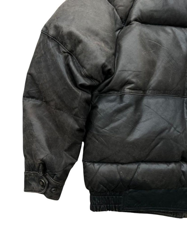 May include: A close-up of a black leather puffer jacket. The jacket has a quilted design and a ribbed hem. The sleeves have buttoned cuffs. The leather appears worn, with a matte finish. The jacket is a classic outerwear piece.