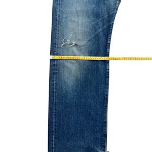 Puede incluir: Vaqueros de mezclilla azul descolorido con una zona de rodilla desgastada. Se colocan cintas m&eacute;tricas amarillas sobre los vaqueros para medir la longitud y el ancho. Los vaqueros est&aacute;n planos sobre una superficie blanca.