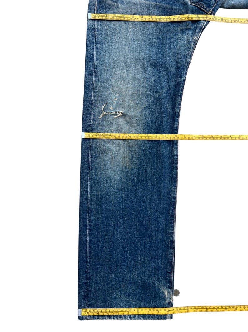 Puede incluir: Vaqueros de mezclilla azul descolorido con una zona de rodilla desgastada. Se colocan cintas m&eacute;tricas amarillas sobre los vaqueros para medir la longitud y el ancho. Los vaqueros est&aacute;n planos sobre una superficie blanca.