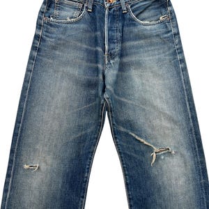 Puede incluir: Vaqueros de mezclilla azul descolorido con bragueta de botones y detalles desgastados. Los vaqueros tienen un dise&ntilde;o cl&aacute;sico de cinco bolsillos y muestran signos de desgaste con rasgaduras en las rodillas. La mezclilla tiene un lavado vintage.