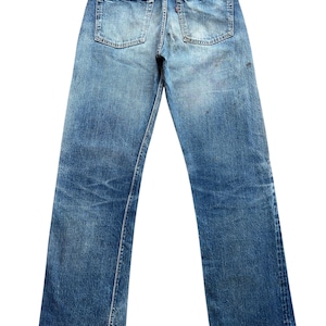 以下が含まれることがあります： 色あせた青色のデニムジーンズ。ダメージ加工が施されており、バックポケットが2つ、革パッチ、裾はほつれています。デニムは使用感と色あせが見られ、ヴィンテージ感のある外観です。