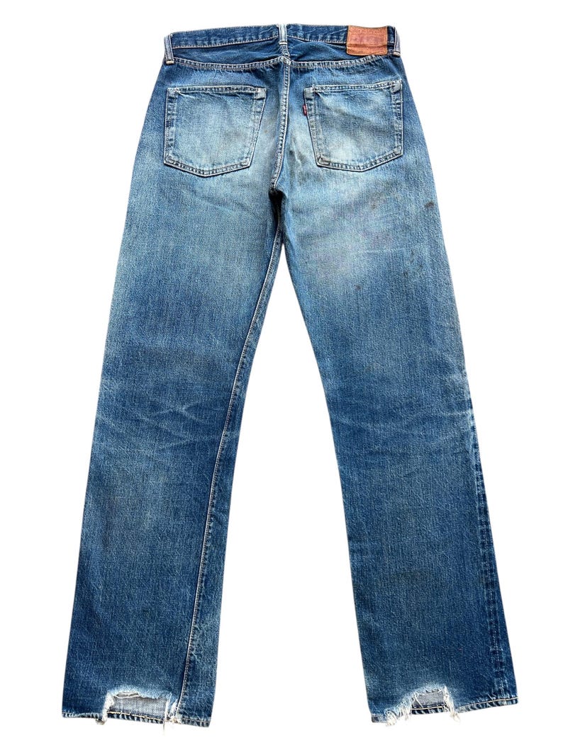 以下が含まれることがあります： 色あせた青色のデニムジーンズ。ダメージ加工が施されており、バックポケットが2つ、革パッチ、裾はほつれています。デニムは使用感と色あせが見られ、ヴィンテージ感のある外観です。