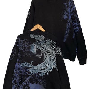 Op de afbeelding: Zwart sweatshirt met een gedetailleerd ontwerp van een feniks en bloemen in blauwe en witte tinten. Het sweatshirt wordt getoond op een houten hanger, waarbij de voor- en achterkant te zien zijn.