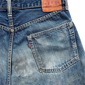 以下が含まれることがあります： 青いデニムジーンズの背面をクローズアップ。ジーンズは、色あせた部分とバックポケットの小さな赤いタグで、使い古された外観をしています。茶色の革のパッチがウエストバンドに見えます。