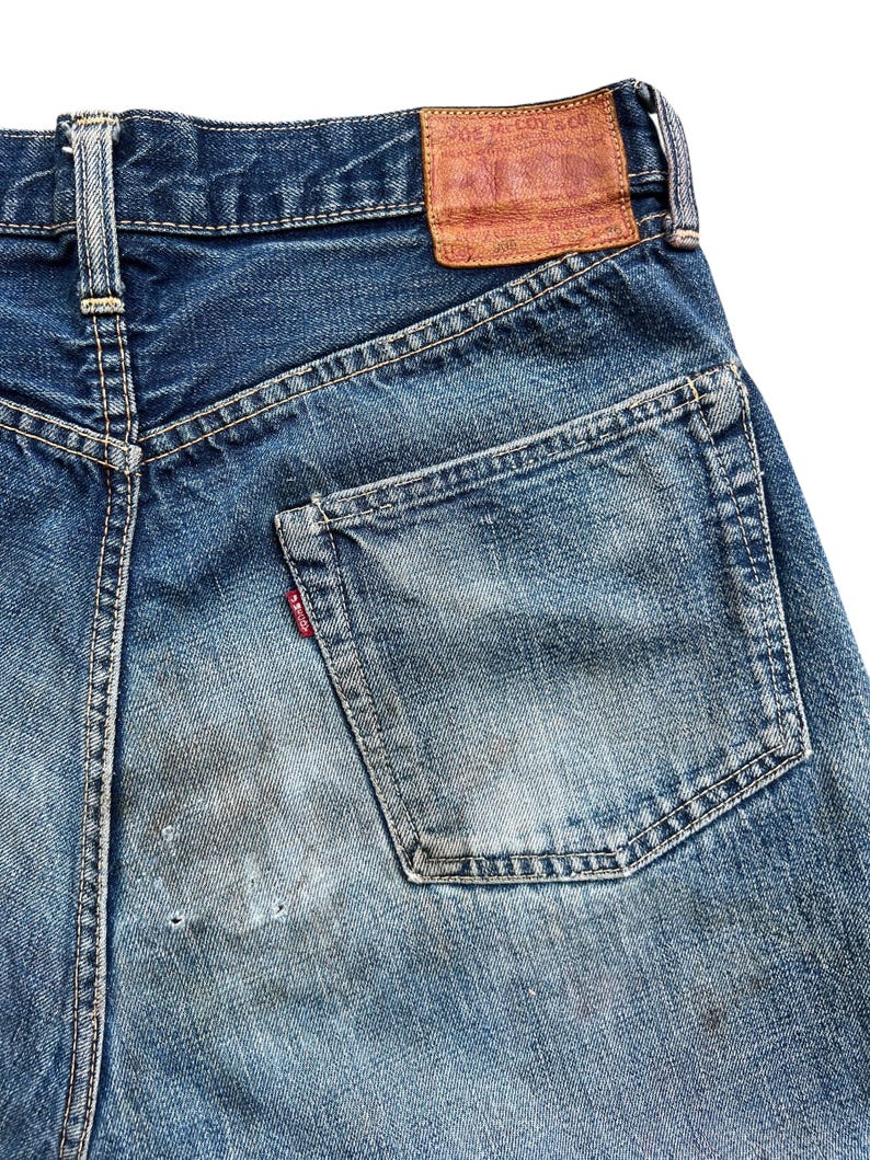 以下が含まれることがあります： 青いデニムジーンズの背面をクローズアップ。ジーンズは、色あせた部分とバックポケットの小さな赤いタグで、使い古された外観をしています。茶色の革のパッチがウエストバンドに見えます。