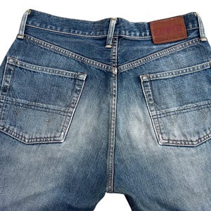 Puede incluir: Primer plano de la parte trasera de unos vaqueros azules. Los vaqueros tienen dos bolsillos traseros y un parche de cuero marr&oacute;n con la marca "EDWIN". El denim tiene un aspecto descolorido y desgastado.