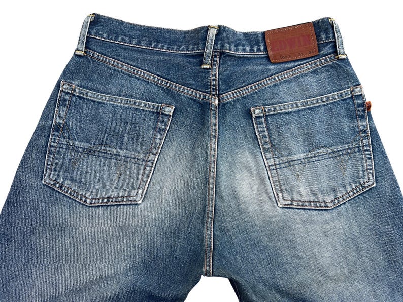 Puede incluir: Primer plano de la parte trasera de unos vaqueros azules. Los vaqueros tienen dos bolsillos traseros y un parche de cuero marr&oacute;n con la marca "EDWIN". El denim tiene un aspecto descolorido y desgastado.