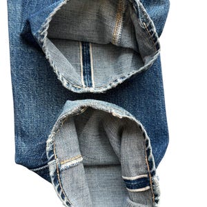 Pu&ograve; includere: Primo piano dell'orlo ripiegato di jeans blu. I risvolti sono ripiegati, rivelando il bordo grezzo con una striscia bianca e blu. Il denim ha un aspetto usurato, con cuciture visibili e un lavaggio sbiadito.
