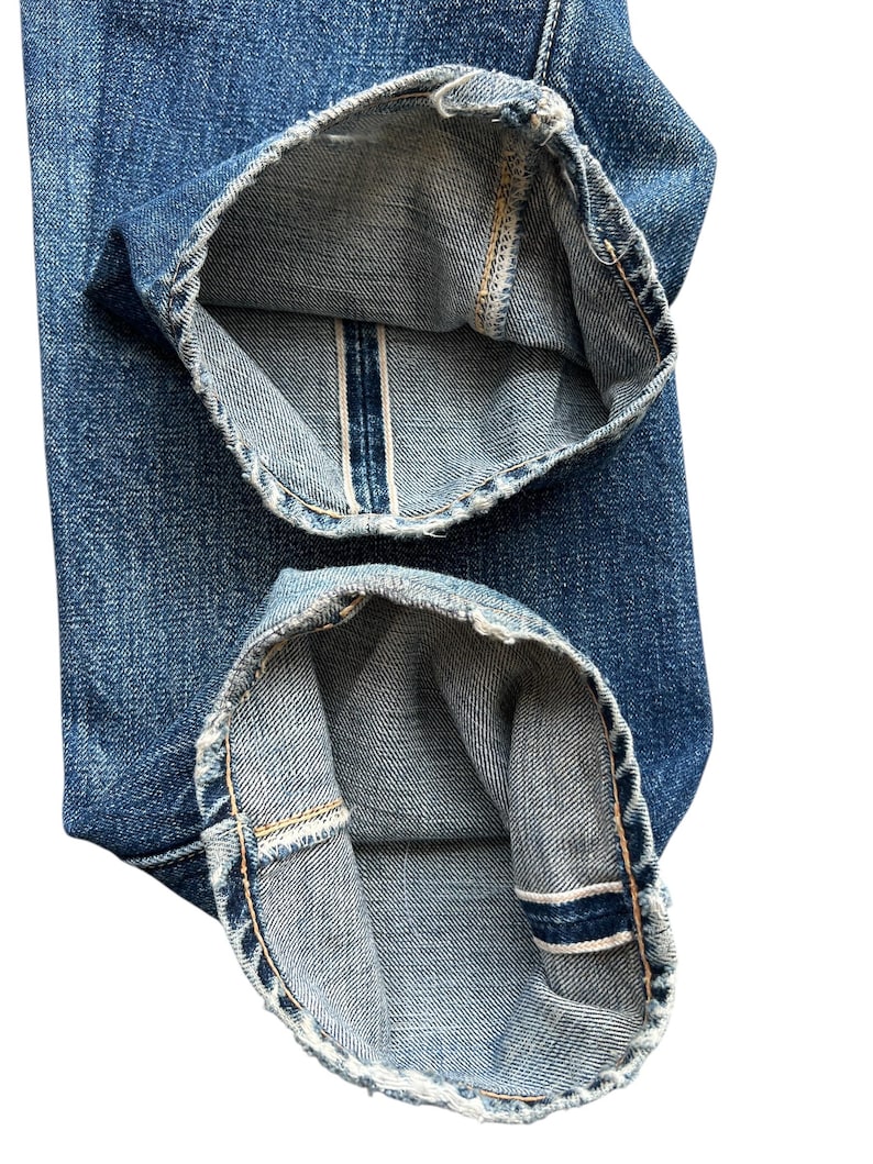 Pu&ograve; includere: Primo piano dell'orlo ripiegato di jeans blu. I risvolti sono ripiegati, rivelando il bordo grezzo con una striscia bianca e blu. Il denim ha un aspetto usurato, con cuciture visibili e un lavaggio sbiadito.