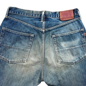 May include: Close-up of the back of blue denim jeans. The jeans have two back pockets, a brown leather patch with text, and a faded, worn appearance. The denim is a medium wash with lighter areas.