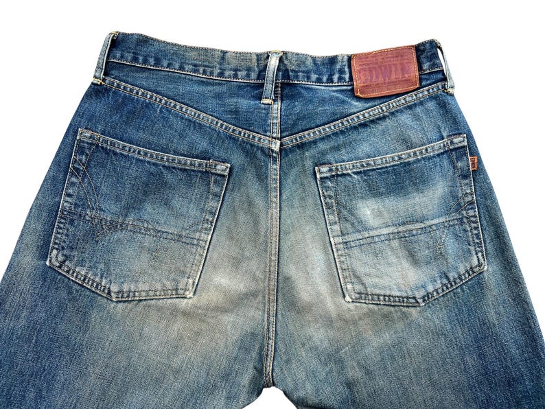 May include: Close-up of the back of blue denim jeans. The jeans have two back pockets, a brown leather patch with text, and a faded, worn appearance. The denim is a medium wash with lighter areas.