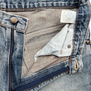K&ouml;nnte beinhalten: Nahaufnahme einer blauen Jeans mit teilweise ge&ouml;ffnetem Rei&szlig;verschluss. Die Jeans hat einen Knopfverschluss mit der Marke "Wrangler". Ein Etikett mit japanischem Text ist ebenfalls sichtbar.