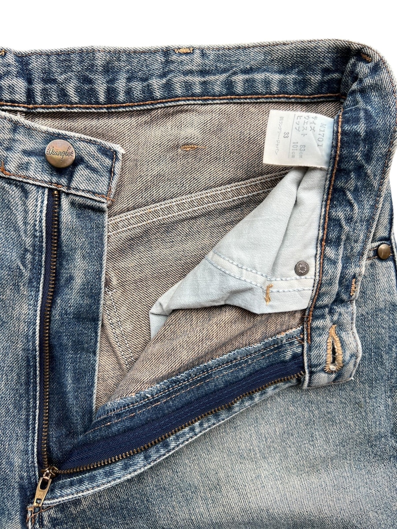 K&ouml;nnte beinhalten: Nahaufnahme einer blauen Jeans mit teilweise ge&ouml;ffnetem Rei&szlig;verschluss. Die Jeans hat einen Knopfverschluss mit der Marke "Wrangler". Ein Etikett mit japanischem Text ist ebenfalls sichtbar.