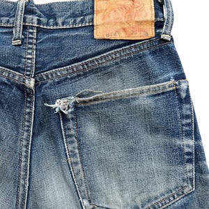 Pu&ograve; includere: Primo piano di jeans di denim blu con un lavaggio sbiadito. La tasca posteriore &egrave; visibile, insieme a una toppa in pelle usurata. I jeans mostrano segni di usura, con un aspetto invecchiato.