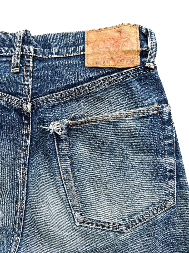 Pu&ograve; includere: Primo piano di jeans di denim blu con un lavaggio sbiadito. La tasca posteriore &egrave; visibile, insieme a una toppa in pelle usurata. I jeans mostrano segni di usura, con un aspetto invecchiato.
