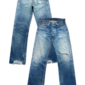 以下が含まれることがあります： ヴィンテージの青いデニムジーンズが2足。1足は後ろから見て、ポケットと革のパッチが見えます。もう1足は前から見て、ボタンフライとダメージ加工の膝が見えます。