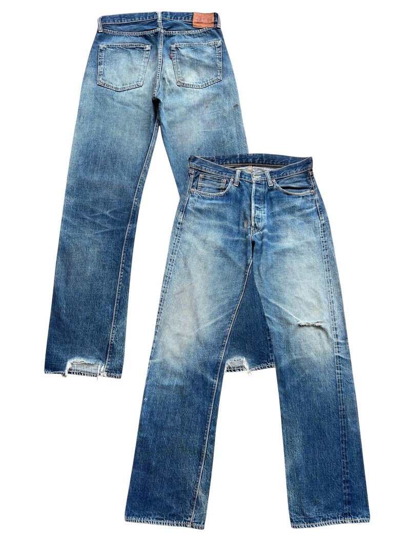 以下が含まれることがあります： ヴィンテージの青いデニムジーンズが2足。1足は後ろから見て、ポケットと革のパッチが見えます。もう1足は前から見て、ボタンフライとダメージ加工の膝が見えます。