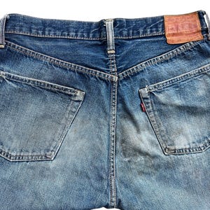 以下が含まれることがあります： 青いデニムジーンズの背面をクローズアップ。ジーンズには2つのバックポケット、右ポケットの小さな赤いタグ、茶色の革パッチが付いています。デニムは色あせ、使い古された外観をしています。